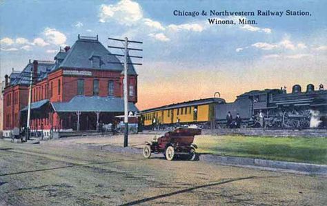 Colorized photo postcard of a red brick train station next to railroad tracks on which a grey steam engine pulling orange cars stands; a violet motorcar, circa 1910, stands on the street outside the station